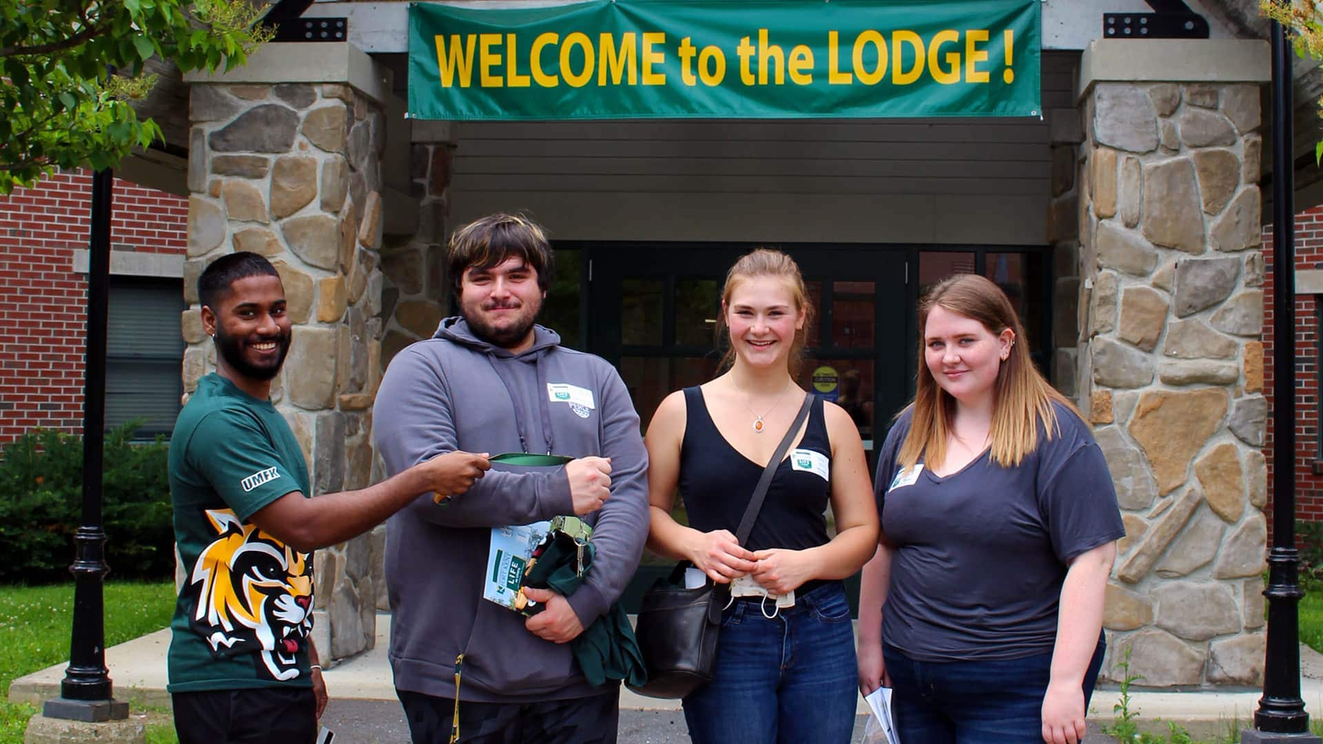 Four students stand together in front of the Lodge entrance. A banner that reads Welcome to the Lodge hangs above the building entryway.