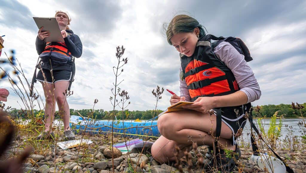 two students gather data by the river with a raft behind them