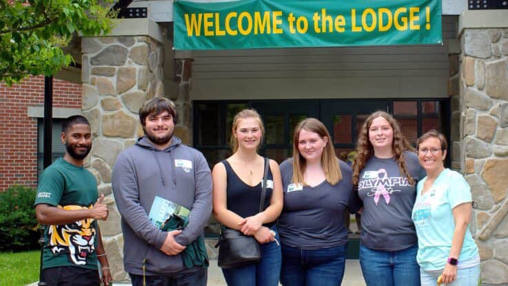 new students, a parent, and a UMFK tour guide pose in front of The Lodge. Above them is a banner over the entryway that says Welcome to The Lodge