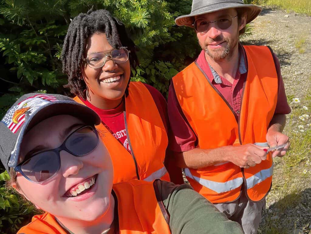 two students and a professor pose in orange vests in front of a tree