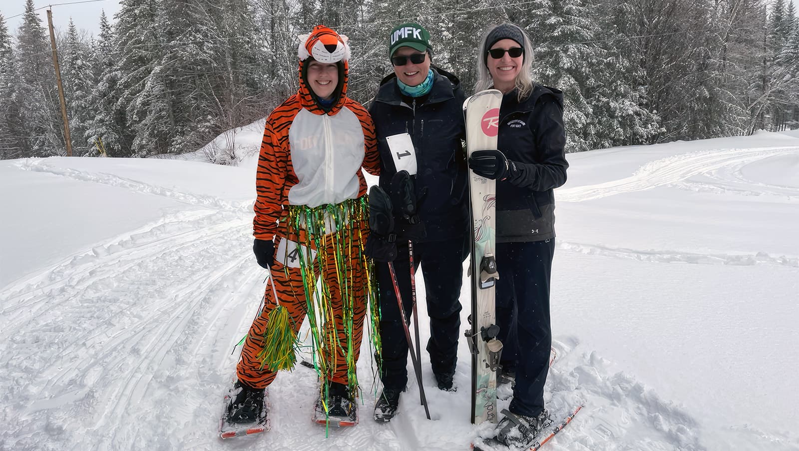 three UMFK employees pose smiling while near the cross-country trail; one of the employees is wearing a bengal costume