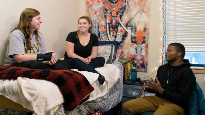 One male and two female students sit in a dorm room talking. The ladies are sitting on a bed while the guy is sitting in a chair.