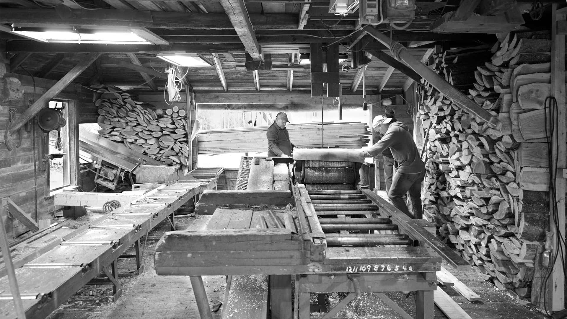 two men stand on opposite sides of a planer they are operating inside a local sawmill
