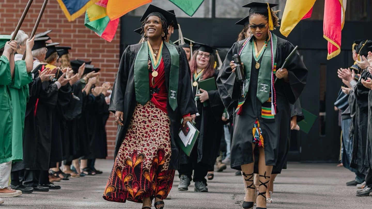 Two female non-traditional students walk out of the Sports Center after their commencement ceremony. One of them, wearing a fiery red dress, smiles as she looks upward with a sense of hope for the future in her eyes