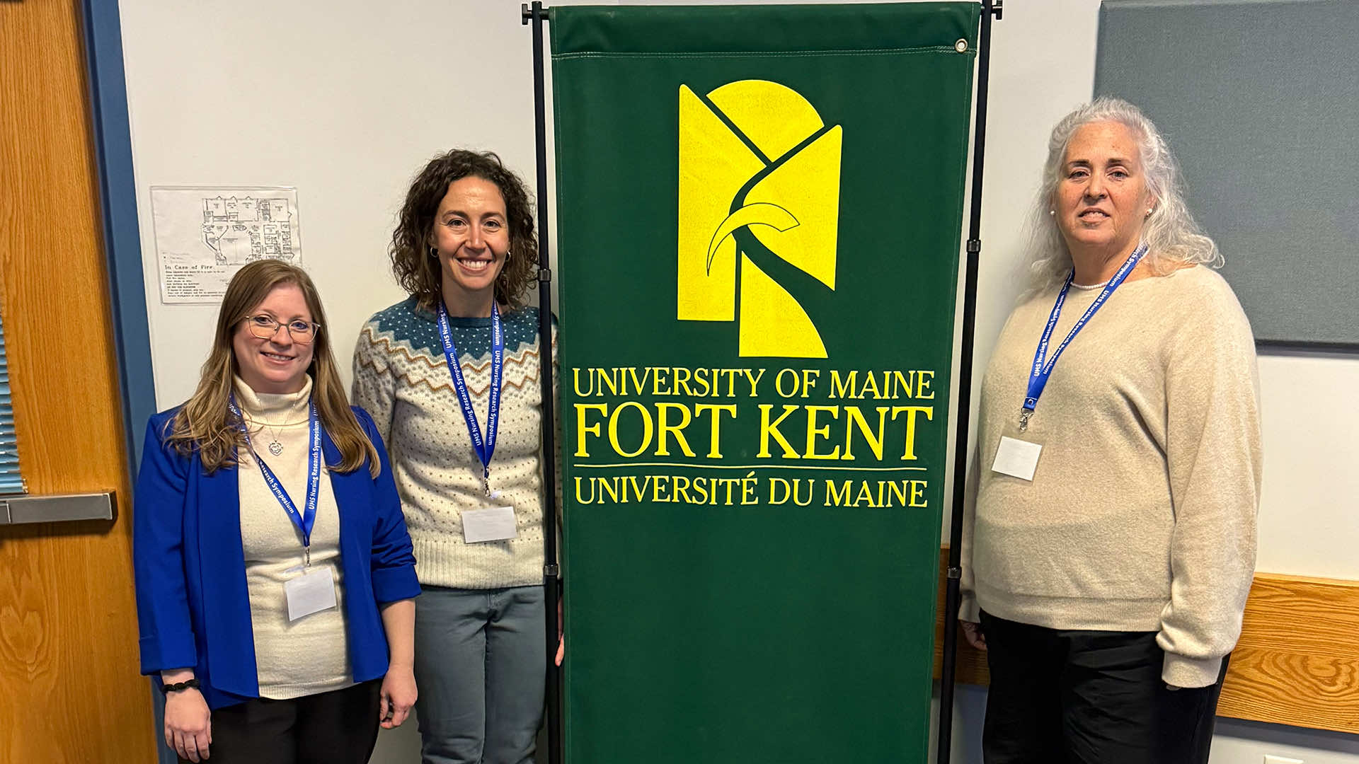 three female faculty members stand beside a University of Maine at Fort Kent banner