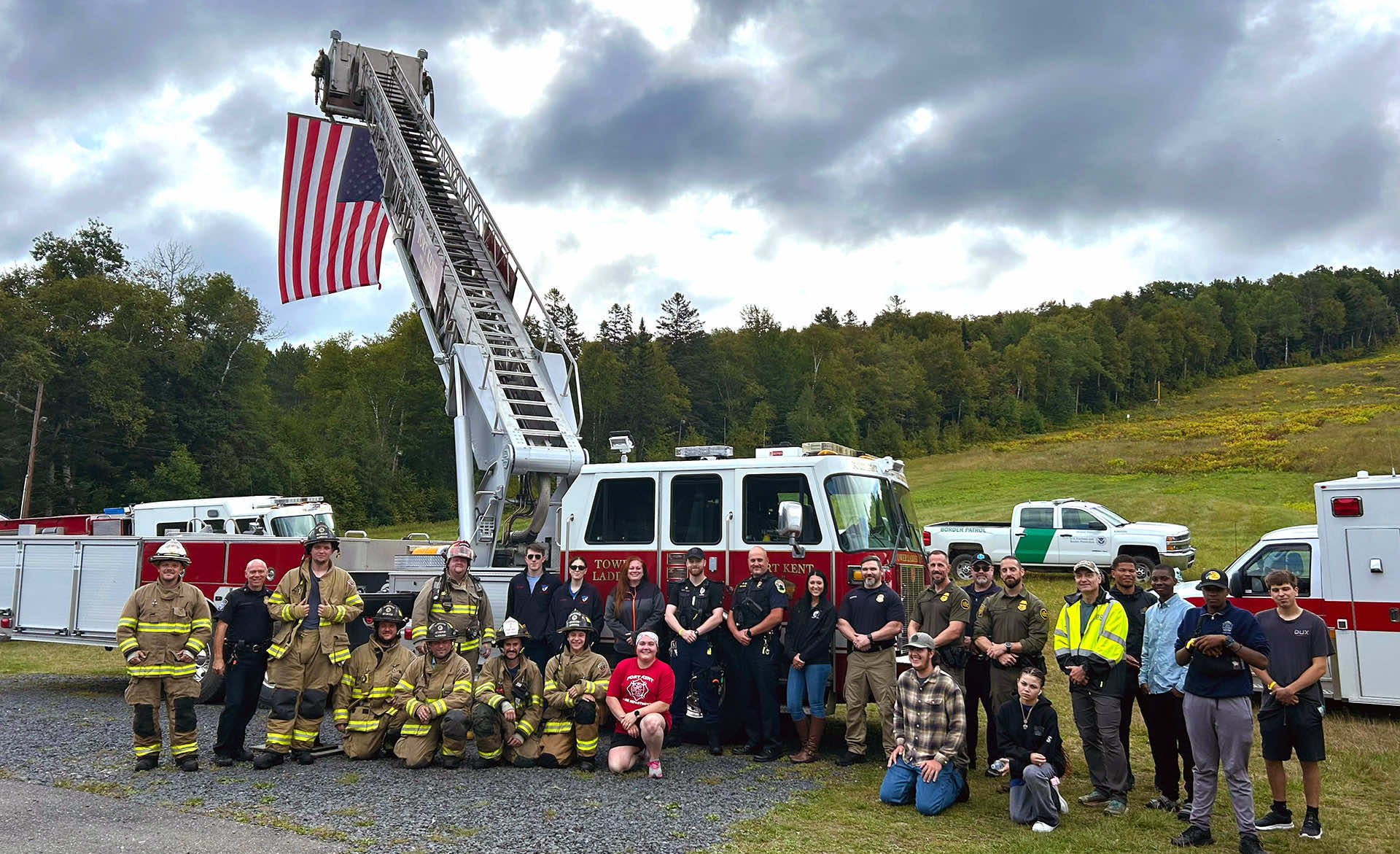 9/11 Memorial Hike 1 first responders pose for a group photo in front of a fire truck, with an American flag hanging from the fire truck's ladder