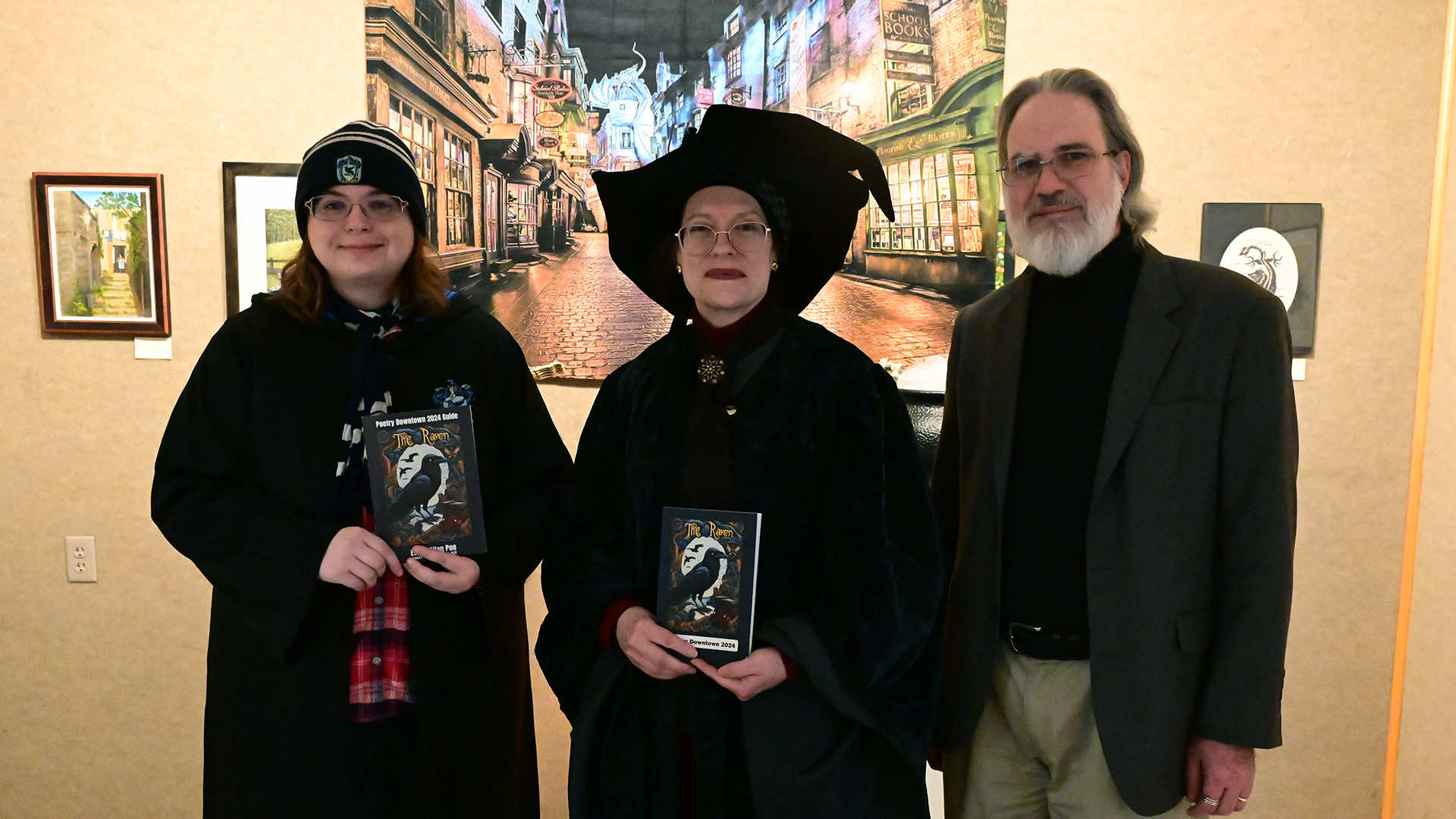 Jessica Boynton, Geraldine Cannon Becker, and Dr. Joseph Becker pose for a group photo in front of some art in the Blake Library Gallery. Jessica and Geraldine are dressed in Harry Potter-inspired costumes, while Joe wears a blazer over a black turtleneck shirt.